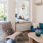 Wohnzimmer mit blauem Sofa, Holztisch und Sessel. Küche im Hintergrund über Glaswand.