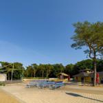 Freiluftbühne mit Bänken, gelbes Gebäude und großer Baum unter blauem Himmel.