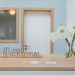 Willkommen-Schild und Blumen auf Holztisch vor Tür und Spiegel.