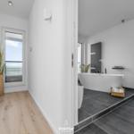 Badezimmer mit freistehender Badewanne und großem Fenster. Holzfußboden und Pflanze im Flur.