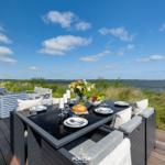Terrasse mit Tisch und Sitzgelegenheiten. Blick auf Wasser und Himmel.