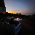 Person sitzt auf Terrasse mit Feuerkorb und Blick auf Wasser bei Sonnenuntergang.