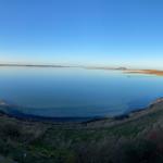 Panoramablick auf ruhiges Wasser und grasbewachsenen Uferbereich bei klarem Himmel.