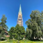 Hoch aufragender Kirchturm mit grünem Zeltdach umgeben von Bäumen und Grünflächen.