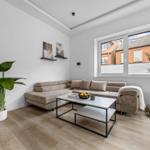 Wohnzimmer mit Ecksofa, Tisch und Pflanzen. Fenster zeigen auf Straßenansicht.