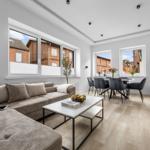 Wohn- und Essbereich mit Sofa, Tisch und Stühlen. Große Fenster mit Blick auf Gebäude.