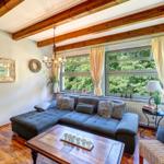 Ein Wohnzimmer mit grauem Sofa, Holzbalken und großem Fenster mit Blick auf Grün.