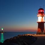 Roter Leuchtturm mit rotem Licht, grüner Leuchtturm im Hintergrund, ruhiges Meer bei Sonnenuntergang.
