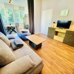 Wohnzimmer mit Sofa, Couchtisch und Fernseher. Durchgang zur Küche mit Tisch und Stühlen.