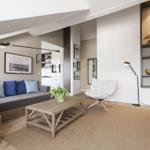 Wohnzimmer mit Sofa, Sessel und Holztisch. Regal mit Dekorationsgegenständen. Küchenbereich im Hintergrund.