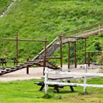 Holzspielplatz mit Rutsche, Klettergerüst und Picknicktischen auf grünem Hang.