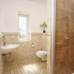 Badezimmer mit Dusche, Waschbecken und Toilette. Fliesen und Fenster sichtbar.