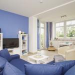 Wohnzimmer mit blauem Sofa, Fernseher und großem Fenster. Essbereich mit Tisch und Stühlen.