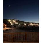 Nachtansicht eines beleuchteten Skigebiets mit Mond am Himmel und Holzgeländer im Vordergrund.