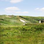 Haus mit weißem Schornstein zwischen grünen Dünen und einem sandigen Pfad.