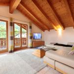 Wohnzimmer mit Holzdecke, Sofa, Fernseher und Blick auf Balkon und Wald.