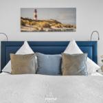 Schlafzimmer mit blauem Kopfteil, weißen Kissen und Wandbild mit Leuchtturm.