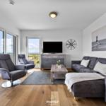 Wohnzimmer mit Sofa, Sesseln und Meerblick über große Fenster.