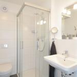 Badezimmer mit Dusche, Waschbecken und Toilette. Weiße Fliesen und graue Handtücher.