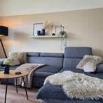 Ein grauer Sofa mit Felldecke und Kissen. Holztisch und Stehlampe daneben.