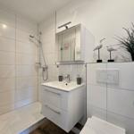 Badezimmer mit Dusche, Waschbecken und Toilette. Weiße Fliesen und Holzboden.