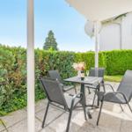 Terrasse mit Tisch, vier Stühlen, Blumen und Weinflasche. Umgeben von grünen Hecken.