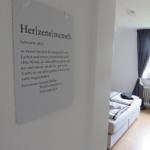 Zimmer mit Bett, Fenster und Wandtafel mit Text.