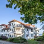 Weißes Mehrfamilienhaus mit Balkonen und rotem Dach unter Bäumen