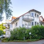 Weißes Mehrfamilienhaus mit Balkonen und rotem Dach unter Bäumen