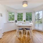 Esszimmer mit Holzboden und Fenstern zum Garten. Tisch und Stühle sind weiß.