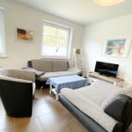 Wohnzimmer mit Sofa, Sessel und Fernseher. Fenster zeigen Wald. Tisch mit kariertem Tischtuch.