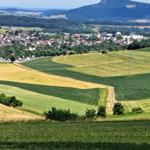 Weitläufige Landschaft mit Feldern, einem Dorf und Bergen im Hintergrund.