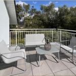 Terrasse mit weißen Sitzmöbeln und Tisch. Blick auf grüne Bäume und einen blauen Himmel.