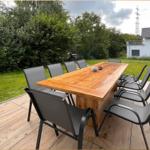 Holztisch mit acht Stühlen auf Terrasse. Grünflächen und Gartenhaus im Hintergrund.