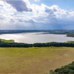 Weitläufiger See mit grünen Wiesen und Waldstreifen unter bewölktem Himmel.