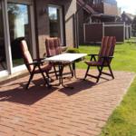 Terrasse mit Tisch und drei Stühlen, umgeben von Gras und Hauswand.