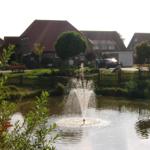 Ein Teich mit Springbrunnen, umgeben von Grün und Häusern im Hintergrund.
