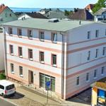 Zweistöckiges Gebäude mit rosa Streifen und Fenstern. Parkplatz und Straße sind sichtbar.