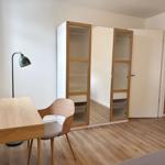 Schreibtisch mit Stuhl und Stehlampe neben Kleiderschrank mit Spiegeltüren.