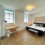 Schlafzimmer mit Bett, Schreibtisch und zwei Fenstern.