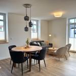 Essbereich mit runder Tisch und Stühlen, Wohnbereich mit Sofa und Sesseln, große Fenster und Holzboden.