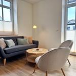 Ein Wohnzimmer mit Sofa, zwei Stühlen und einem runden Tisch. Große Fenster mit Jalousien.