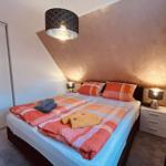 Schlafzimmer mit Bett, Nachttisch und Kleiderschrank. Decke und Kissen sind rot-orange kariert.