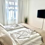 Schlafzimmer mit Bett, TV und Fenster mit weißen Vorhängen.