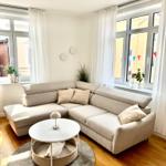 Wohnzimmer mit Ecksofa, runden Tisch und Holzboden. Große Fenster mit weißen Vorhängen.