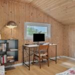 Schreibtisch mit Monitor, Stuhl und Bücherregal an Holzwand. Fenster zeigt Außenansicht.