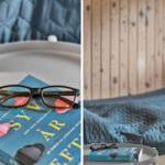 Ein Buch und eine Brille auf einem Tisch neben einem Bett mit blauer Bettdecke.