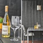 Ein Tisch mit Weinflasche und Gläsern auf einer Terrasse mit dunkler Holzwand.