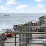 Meerblick mit Segelyacht, Parkplatz und Holzterrasse mit Metallgeländer.