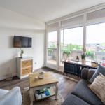 Wohnzimmer mit Sofa, Couchtisch und TV. Balkon mit Pflanzen und Blick auf Gebäude.